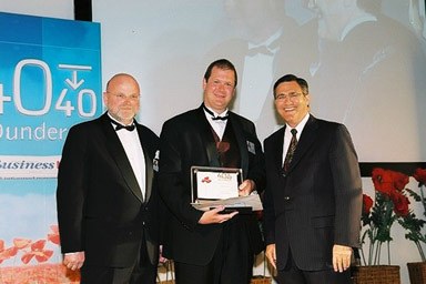 Nathan Buzza accepting the 40 Under 40 First Amongst Equals plaque on stage at the 2003 WA Business News awards, flanked by event hosts.