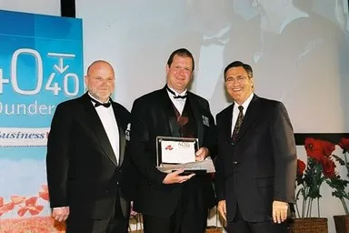 Nathan Buzza accepting the 40 Under 40 First Amongst Equals plaque on stage at the 2003 WA Business News awards, flanked by event hosts.