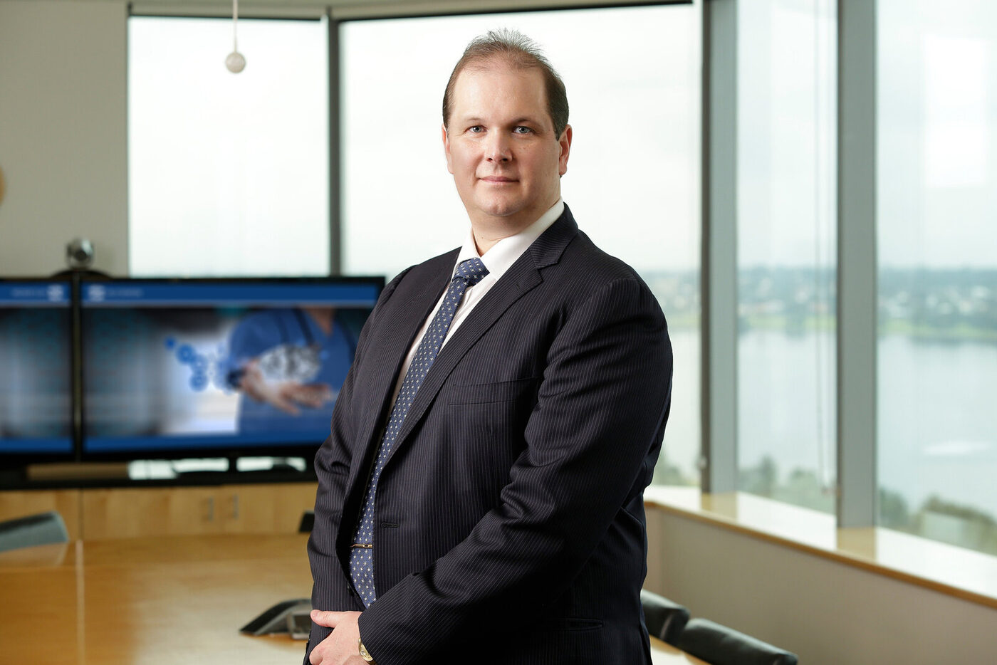 Nathan Buzza in the Alcidion boardroom &mdash; the Miya clinical-imaging display visible behind him, Swan River through the floor-to-ceiling windows.