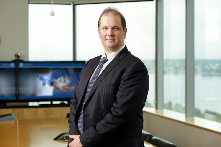 Nathan Buzza in the Alcidion boardroom &mdash; the Miya clinical-imaging display visible behind him, Swan River through the floor-to-ceiling windows.